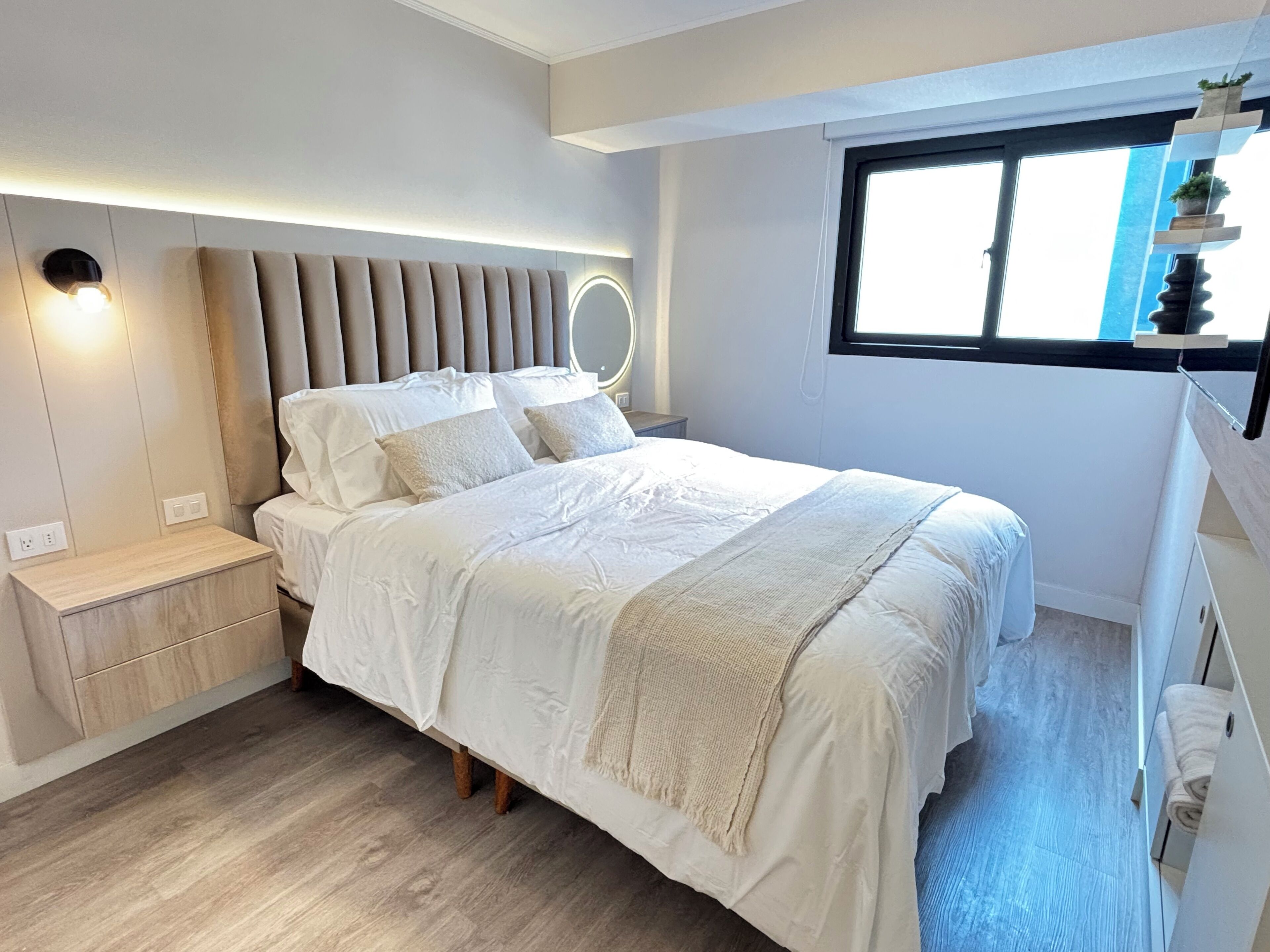 A modern bedroom with a bed, bedside tables, a built-in headboard with lighting, and a window with a view.