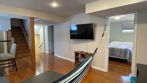 A spacious living area with a wooden floor, a TV mounted on the wall, a hammock, and a bed in the adjacent room.