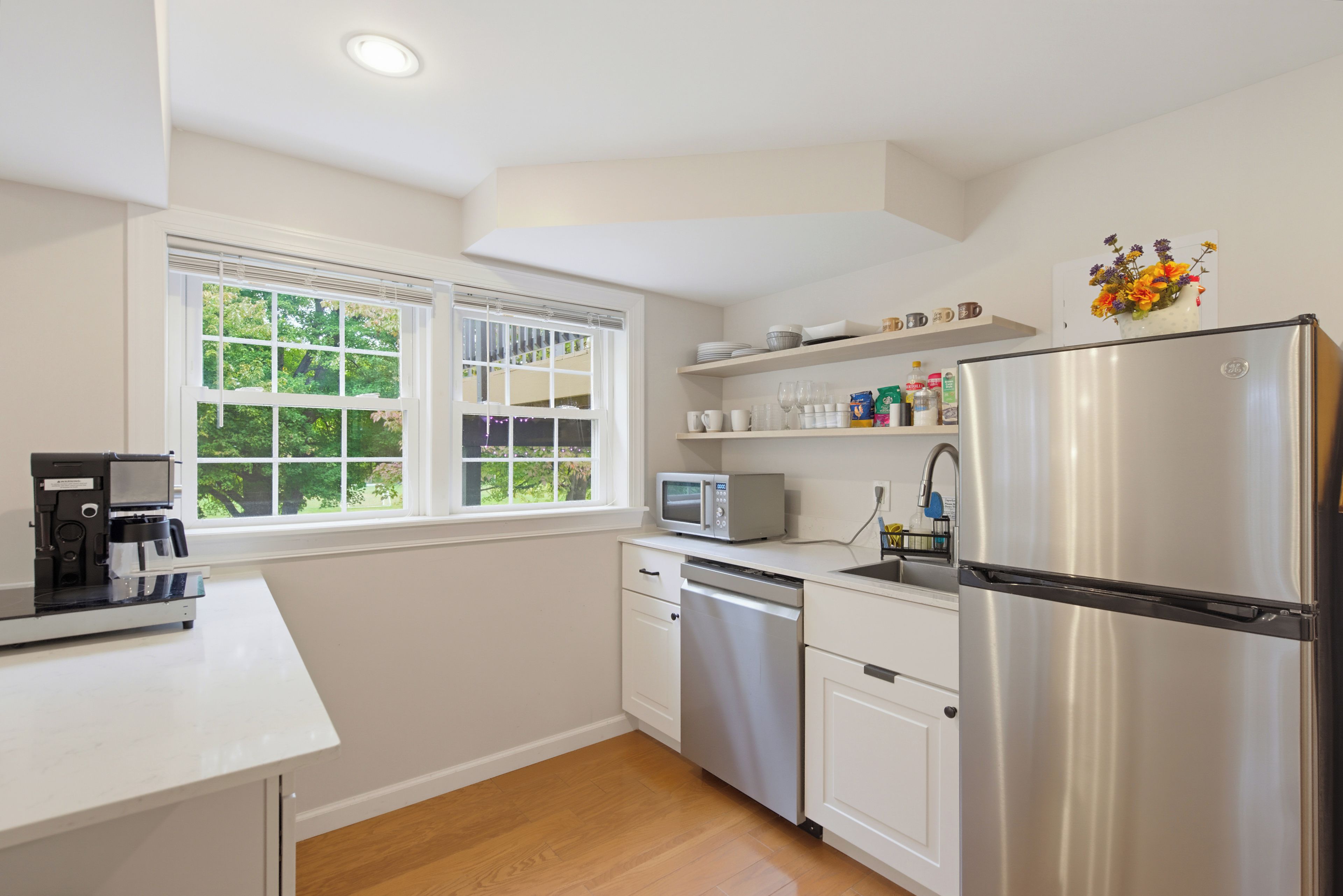 A modern kitchen with white cabinetry, a stainless steel refrigerator, a coffee maker, and a window with a view of greenery.