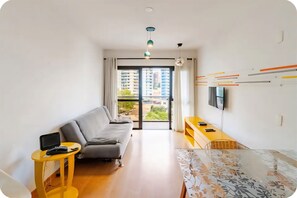 Ein modernes Wohnzimmer mit einer Couch, einem kleinen Tisch, einem an der Wand befestigten Fernseher und einem Balkon mit Blick auf Gebäude.