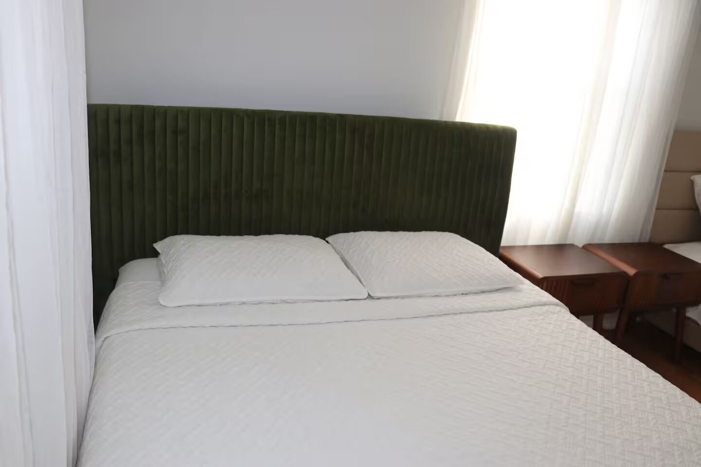 Un lit avec une tête de lit rembourrée de tissu vert, une literie blanche et une table de chevet en bois avec une lampe.