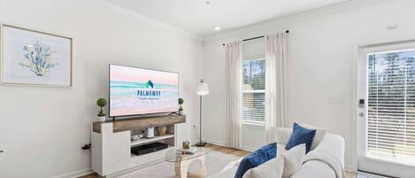A modern living room with a flat-screen TV, a sofa, a coffee table, and a large window with blinds.