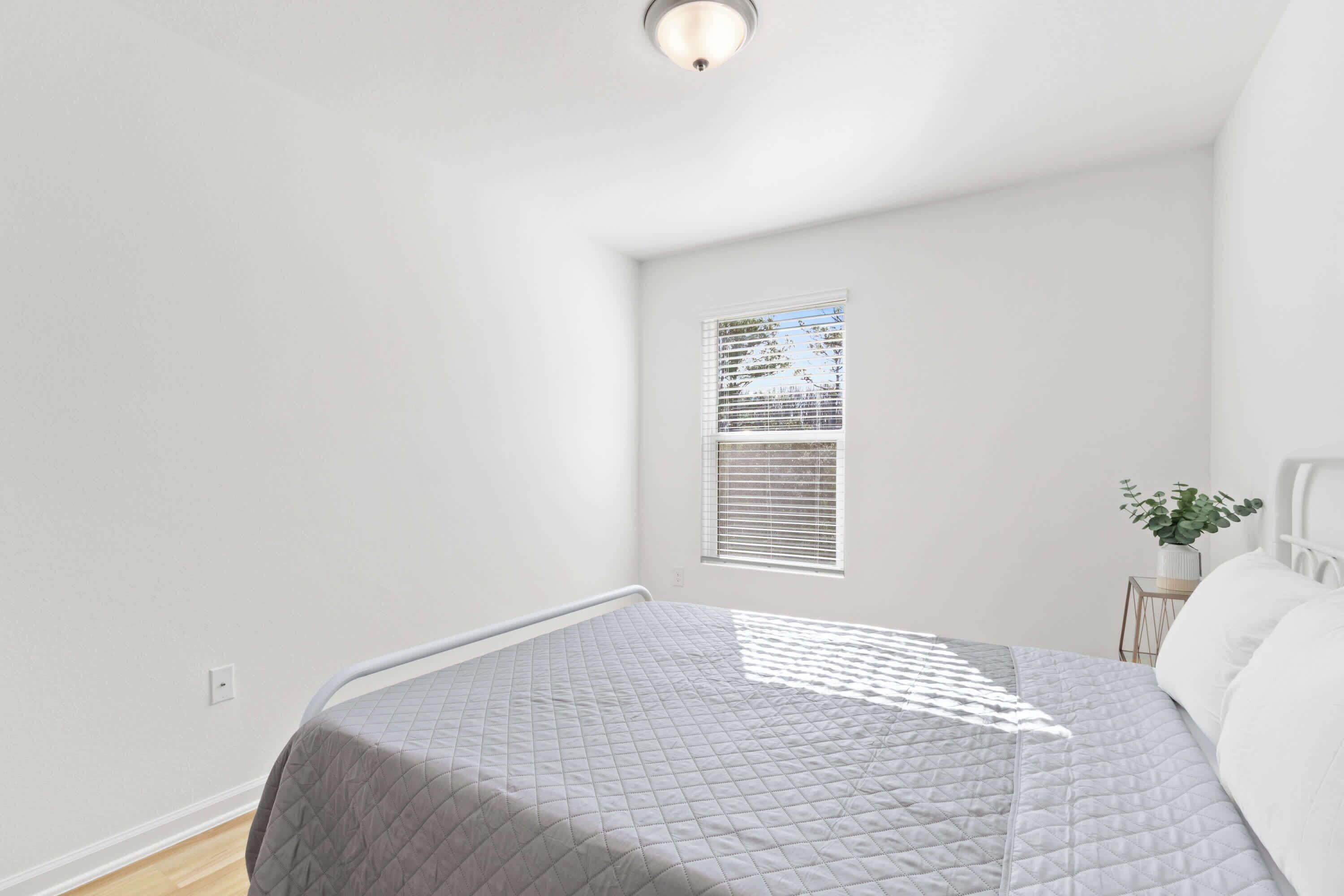 Een slaapkamer met een bed, een raam met zonwering, een klein tafeltje met een plant en een witte plafondlamp.