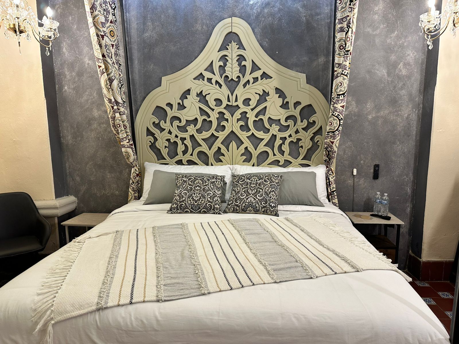 Una cama con un cabecero decorativo, una mesita de noche con un control remoto y una silla.