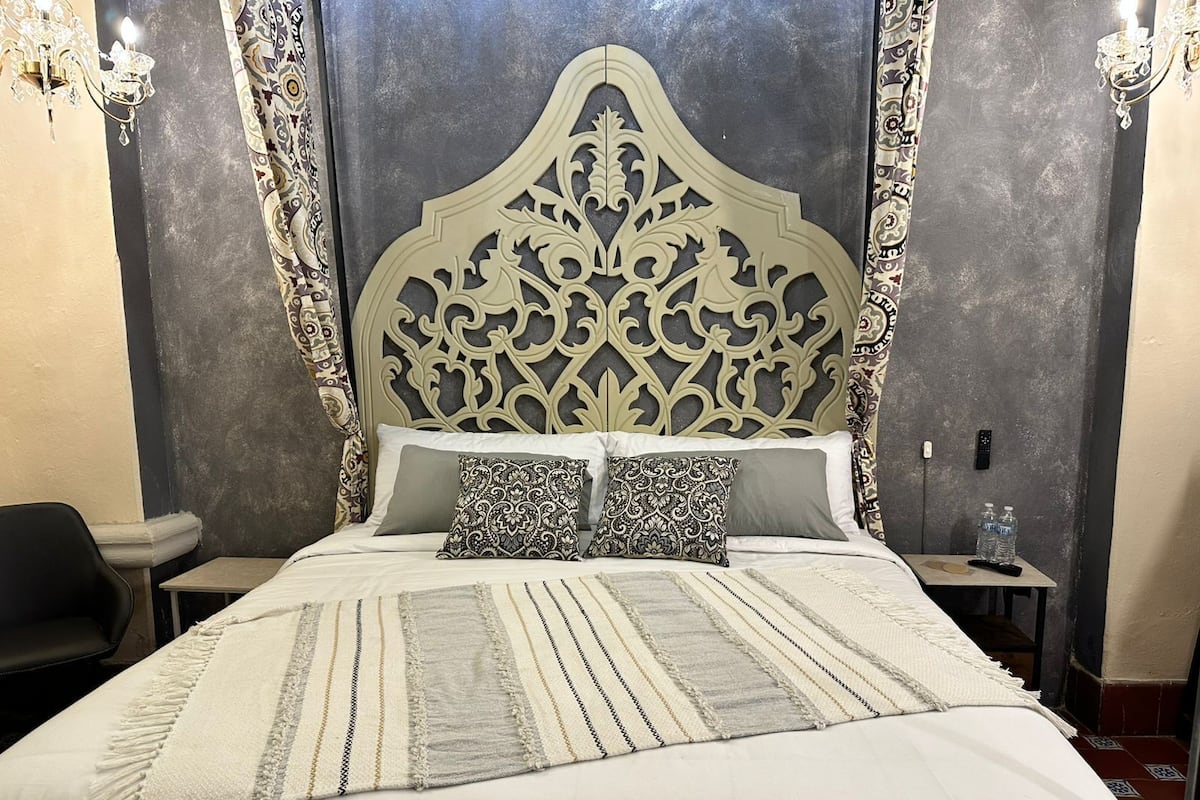Una cama con un cabecero decorativo, una mesita de noche con un control remoto y una silla.