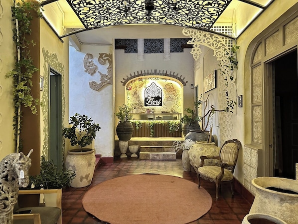 Un interior con un mostrador de recepción, plantas en macetas, un tapete estampado y decoraciones de pared ornamentadas.