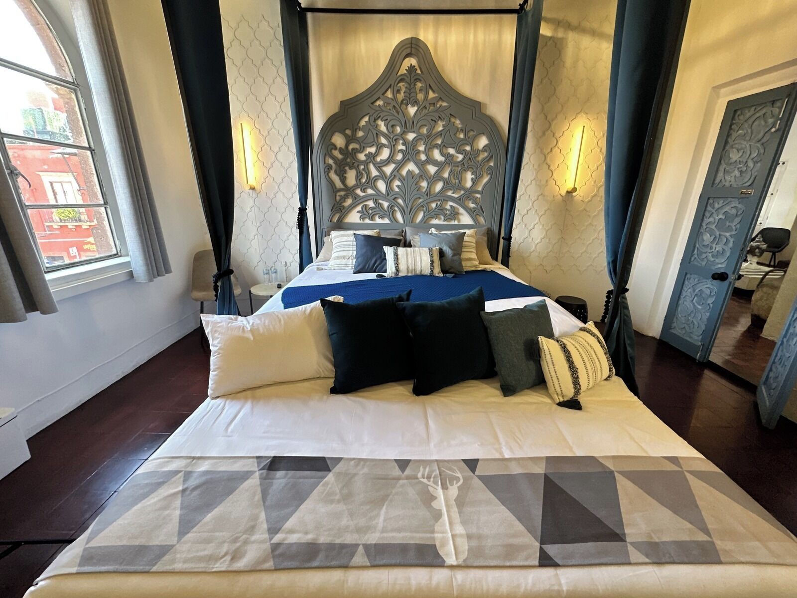 Una cama con dosel con un edredón estampado con motivos geométricos, un cabecero decorativo y dos lámparas fijadas a la pared.