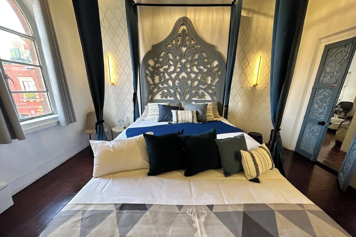 Una cama con dosel con un edredón estampado con motivos geométricos, un cabecero decorativo y dos lámparas fijadas a la pared.