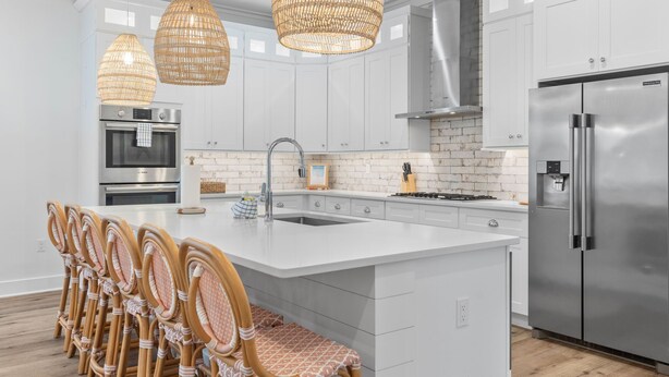 Una cocina moderna con armarios blancos, electrodomésticos de acero inoxidable, lámparas colgantes de mimbre y una isla central con taburetes de bar.