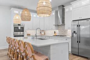 Une cuisine moderne avec des meubles blancs, des appareils en acier inoxydable, des suspensions en osier et un îlot central doté de tabourets de bar.