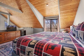 Un dormitorio con techo inclinado, una cama grande con colcha estampada, un aparador, un televisor sobre un mueble bajo y una ventana con cortinas.