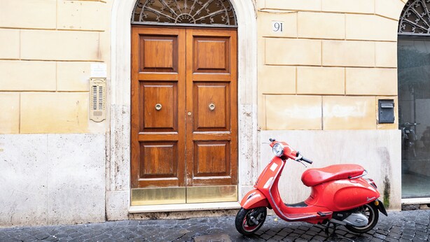 Un scooter rouge est garé devant un immeuble dont la porte est en bois et sur laquelle on peut voir le numéro 91.