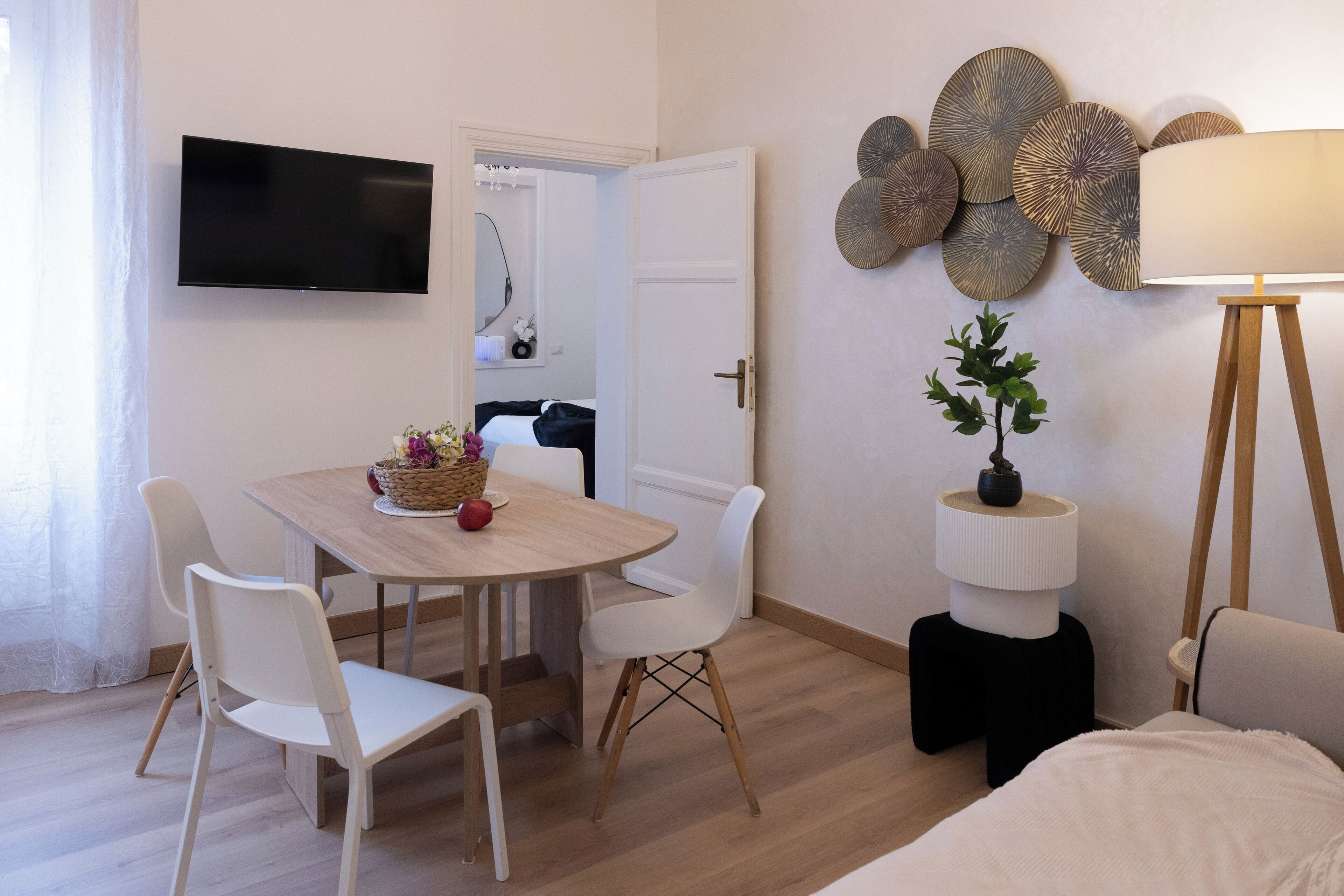 A modern room with a dining table, white chairs, a TV mounted on the wall, decorative wall art, and a lamp.