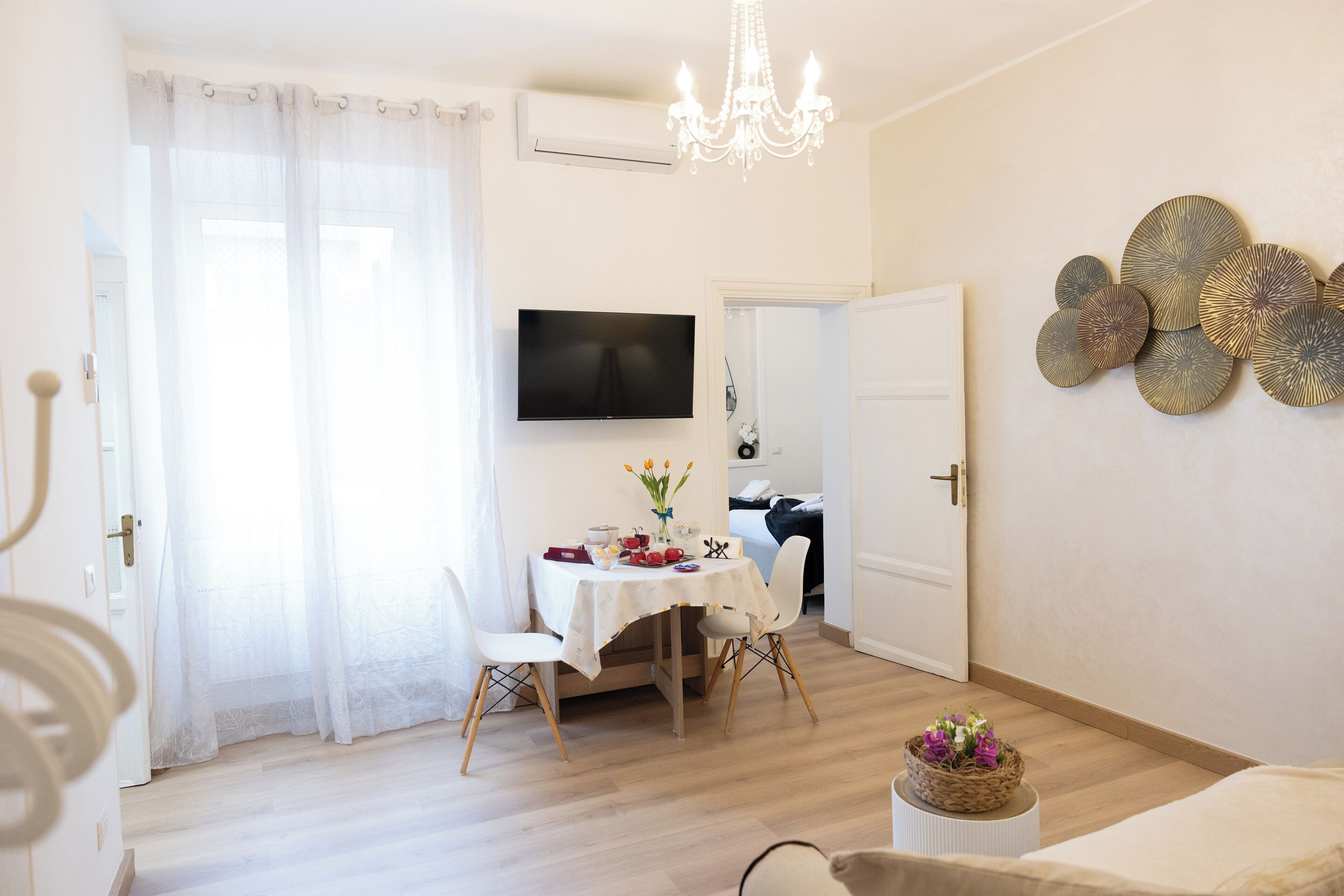 A bright living room with a dining area, a chandelier, a flat-screen TV, and a decorative wall piece.