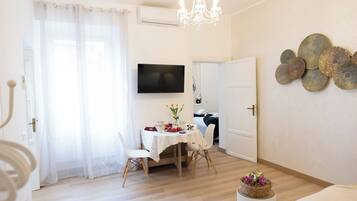 A bright living room with a dining area, a chandelier, a flat-screen TV, and a decorative wall piece.