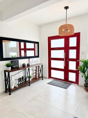 A modern entryway with a red door, a console table with decorative items, a large mirror, and a hanging light fixture. - Casa Marty. Spacious 4-bedroom house in pleasant Aguadilla with cool AC breeze. (Aguadilla)