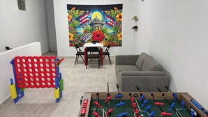 Ein Zimmer mit einem Kicker, einem Sofa, einem kleinen Tisch mit Stühlen und einer farbenfrohen Wandteppich.
