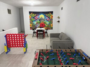 A room with a foosball table, a sofa, a small table with chairs, and a colorful tapestry on the wall. - Casa Marty. Spacious 4-bedroom house in pleasant Aguadilla with cool AC breeze. (Aguadilla)