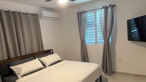 Ein Schlafzimmer mit Bett, Vorhängen, einer Klimaanlage, einem Fenster mit Jalousien und einem an der Wand befestigten Fernseher.