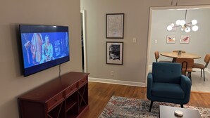 A living room with a flat-screen TV, a wooden entertainment unit, a blue armchair, a dining table with chairs, and a patterned rug. - Fabulous 3-bedroom MidMod Cottage in charming downtown Cary. (Cary)