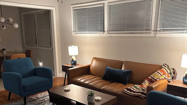 A living room with a brown leather sofa, two blue armchairs, a coffee table, and a patterned rug. There is a ceiling fan, a window with blinds, and a small side table with a lamp. - Fabulous 3-bedroom MidMod Cottage in charming downtown Cary. (Cary)