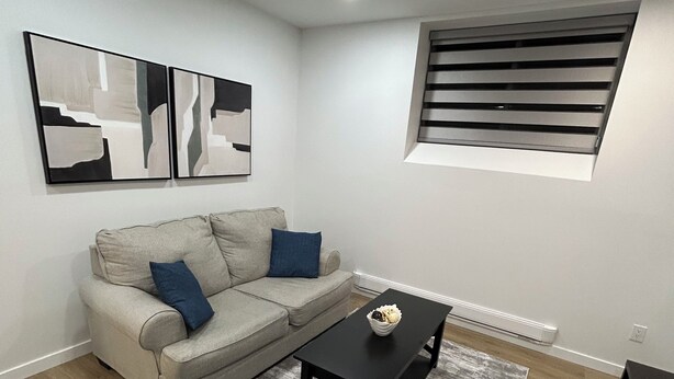A modern living room with a grey sectional sofa, a black coffee table, and a grey armchair. There are abstract wall art pieces and a window with blinds. - Modern Basement Suite in trendy location in South Winnipeg, near UManitoba (Winnipeg)