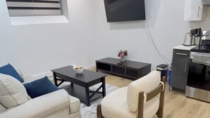 A living room with a sofa, a coffee table, a TV mounted on the wall, and a kitchen area with a stove and cabinets. - Modern Basement Suite in trendy location in South Winnipeg, near UManitoba (Winnipeg)
