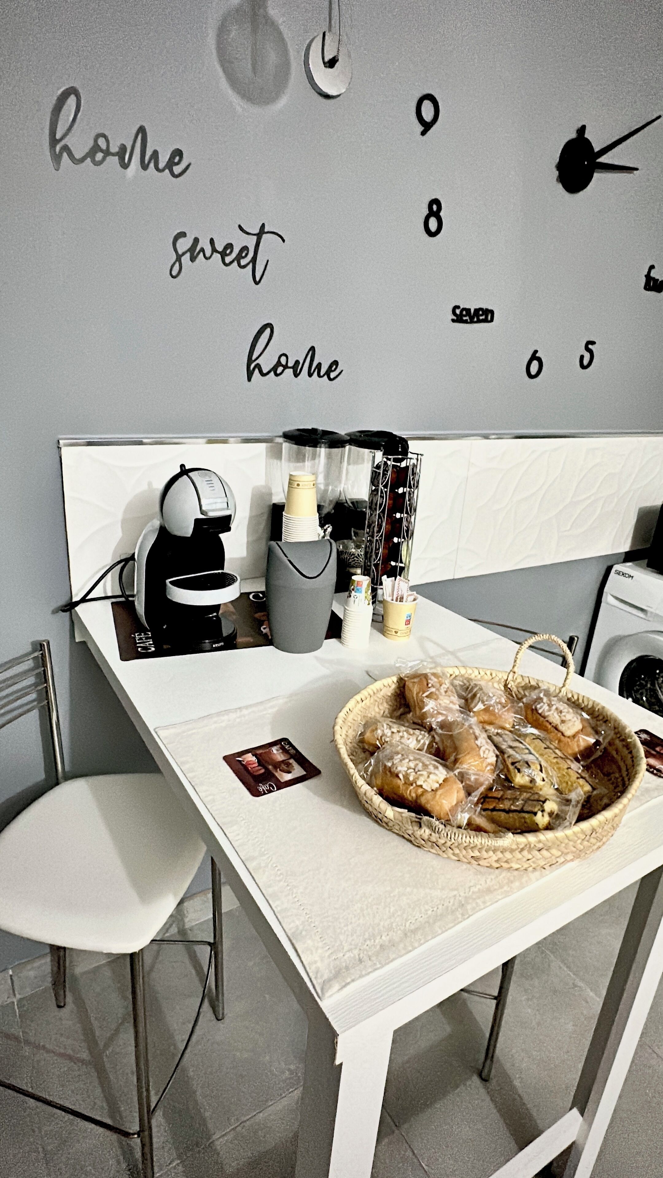 A modern kitchen with a white countertop, a coffee machine, a basket of bread, and a wall clock.