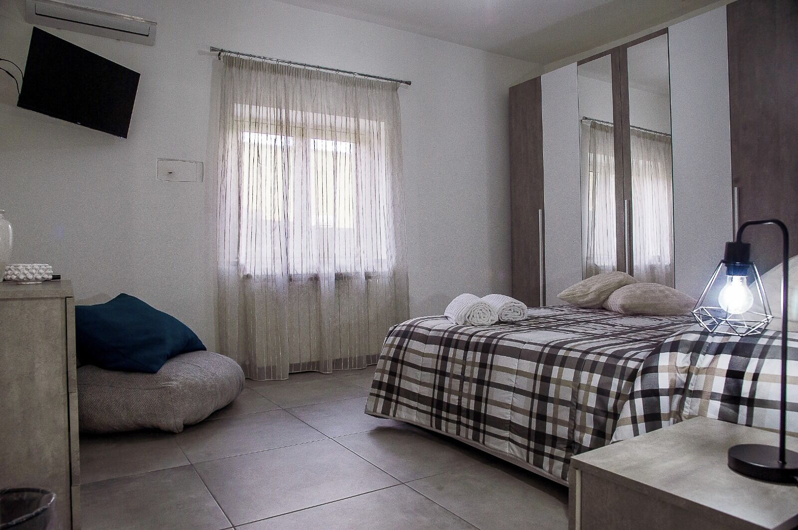 A bedroom with a bed, a bedside table, a lamp, a mirror, a window with curtains, a sofa, and a TV on the wall.