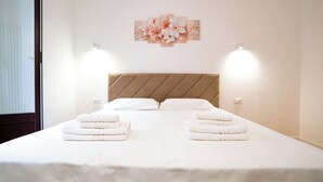 Una cama bien hecha con ropa blanca y dos toallas dobladas encima. Se aprecia un cabecero con diseño diagonal, y dos lámparas fijadas a la pared.