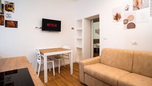 Una sala moderna con una mesa de madera, sillas blancas, un sofá color beige y un televisor instalado en la pared mostrando 'NETFLIX'.