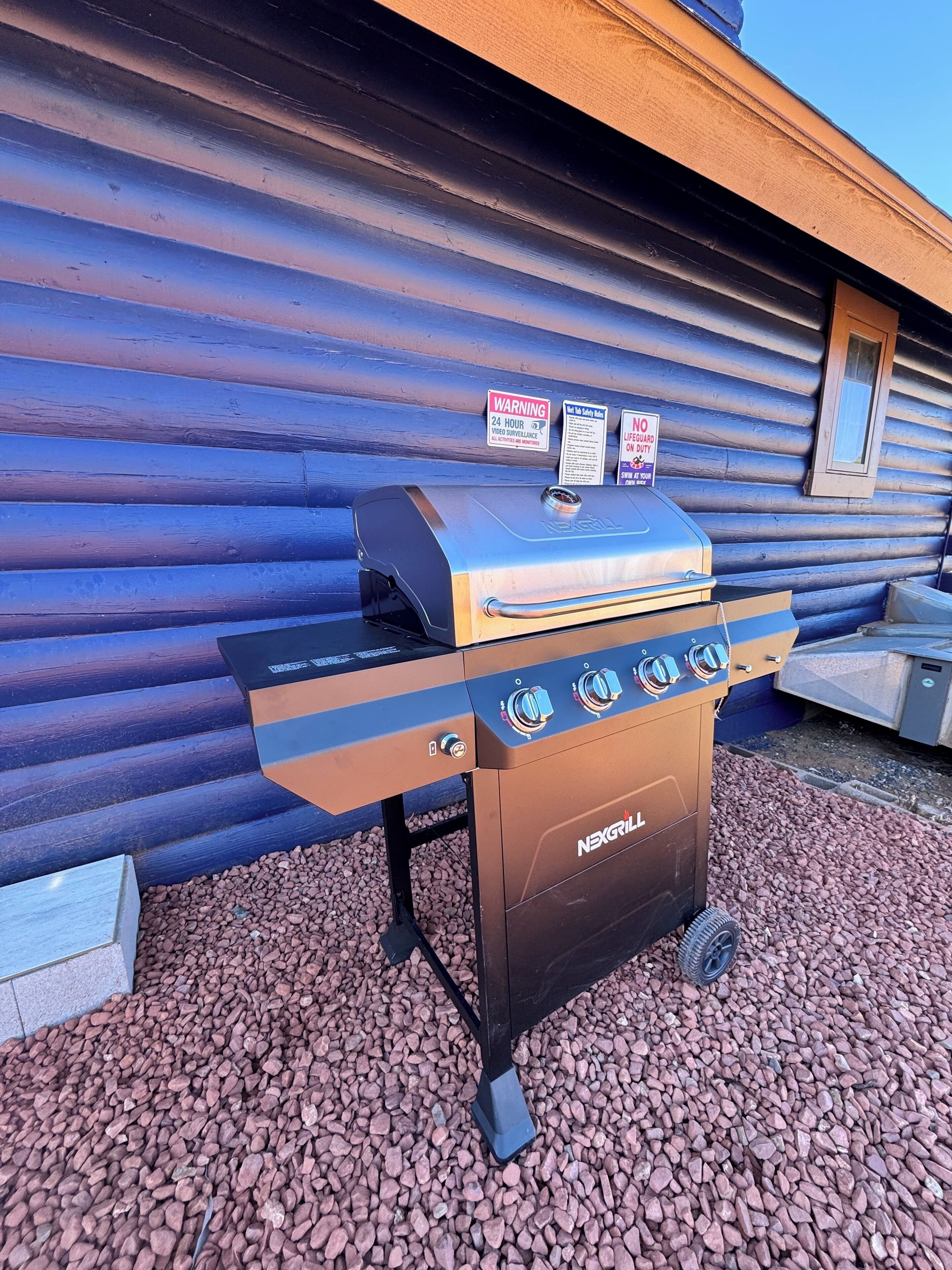 Un barbecue NEXGRILL doté d’un écran numérique et de plusieurs boutons de contrôle, placé sur une surface de gravier devant un bâtiment en bois.