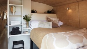 Ein Schlafzimmer mit einem Bett, einer weißen Regalkonstruktion mit gefalteten Handtüchern, einem kleinen Hocker und einem Nachttisch mit Spiegel und Uhr.