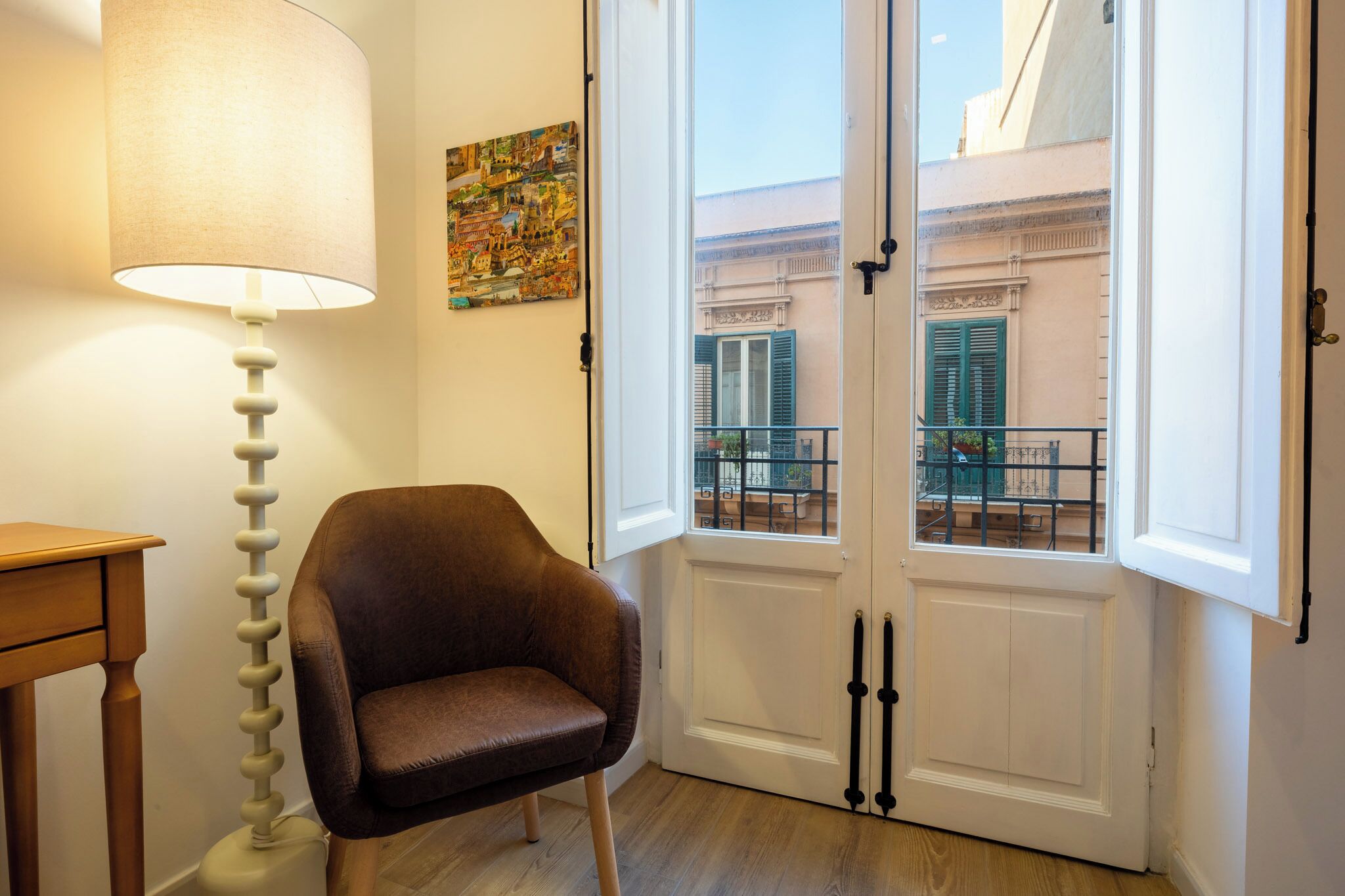 Una habitación con una butaca de cuero marrón, una lámpara de pie blanca, una puerta blanca con vistas al balcón y un cuadro en la pared.