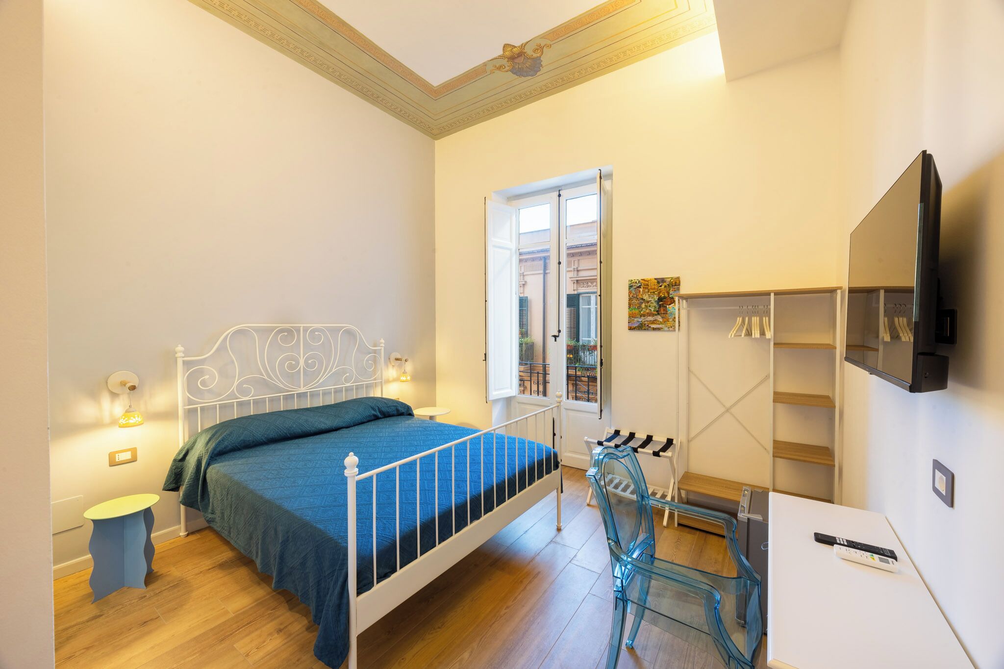 Un dormitorio con cama, mesita de noche, lámpara, ventana y televisor montado en la pared.
