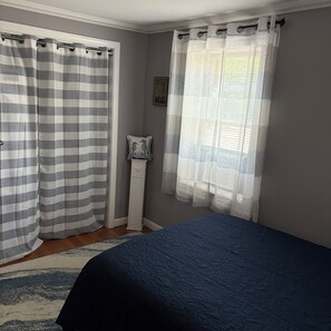 Un dormitorio con cama, una ventana con cortinas de gasa, una cortina a rayas, una mesita de noche blanca con un cojín decorativo y una alfombra estampada.