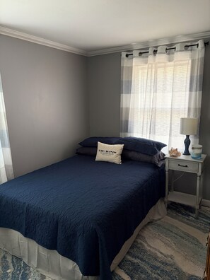 Un dormitorio con cama, mesita de noche con lámpara, ventana con cortinas y almohada con un logotipo.