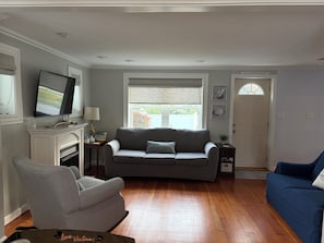 A living room with a grey sofa, a blue sofa, a striped armchair, a fireplace, a television, and a door labeled 'MARSHFIELD'. - Charming 3BR/1BA home short walk to beach fully equipped for relaxing vacation (Marshfield)