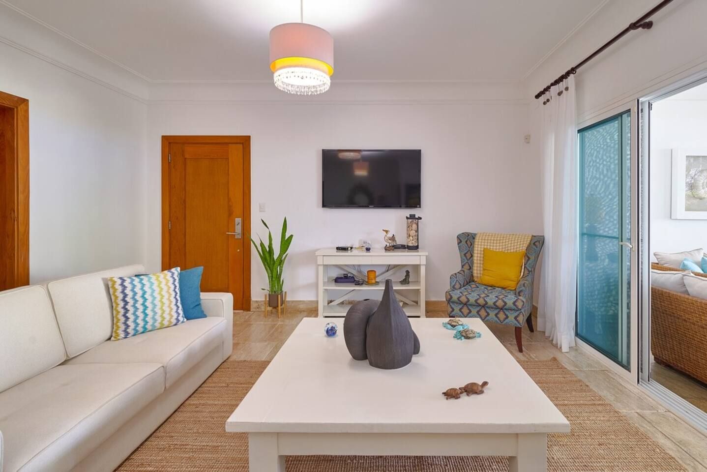 Una sala de estar con un sofá blanco, una mesa de centro, un televisor y un sillón con cojín.