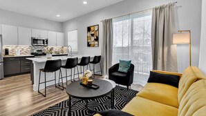 Uma sala de estar moderna com um sofá amarelo, uma área de jantar com uma mesa redonda e uma cozinha com armários brancos e uma janela grande.