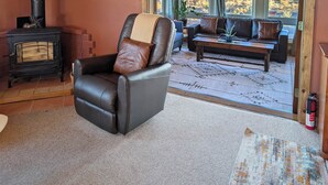 A living room with a leather chair, a sofa, a fireplace, and a view of the desert through large windows. - Bluebird Trail Retreat: Off-Grid & Dark Skies (Saguache County)