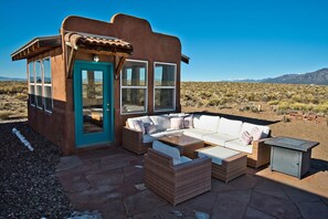 A small, rustic building with a turquoise door and large windows, furnished with a white sofa and wicker chairs, set against a desert landscape.