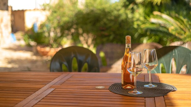 A wooden table with a bottle and two glasses on a placemat, set outdoors with chairs and greenery in the background. - Le due palme - Three Bedroom Apartment, Sleeps 6 (La Caletta)