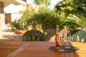 Une table en bois avec une bouteille et deux verres posés sur un set de table, installée à l’extérieur, avec des chaises et de la verdure en arrière-plan.