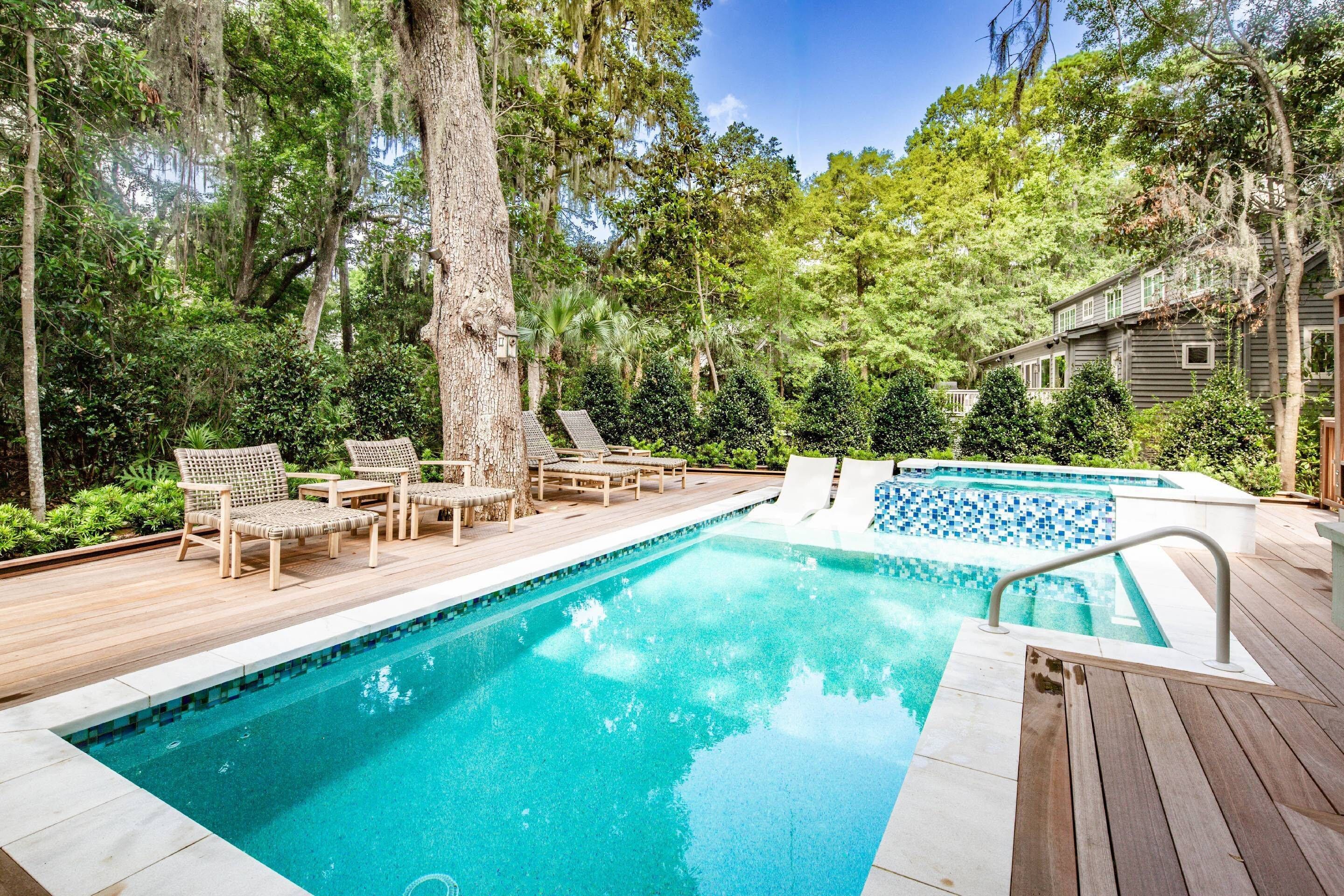 A swimming pool with lounge chairs and a wooden deck, surrounded by trees and a house.