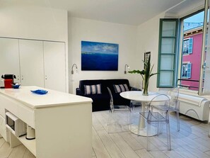 Una cocina moderna con armarios blancos, un sofá negro y una zona de comedor con sillas traslúcidas.