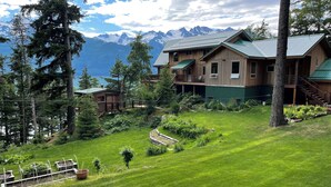 A wooden cabin with a green roof, surrounded by lush greenery and a view of snow-capped mountains.