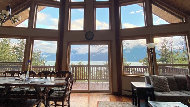 A spacious room with a high ceiling and large windows, a dining area with wooden furniture, and a view of the outdoors.