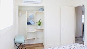 Une chambre à coucher comprenant un lit, une chaise, une armoire avec des étagères, une plante en pot et un appareil de climatisation.