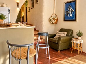 Un espacio de sala acogedor con un sofá verde, un sillón de mimbre, una mesita redonda y una zona de bar con taburetes.
